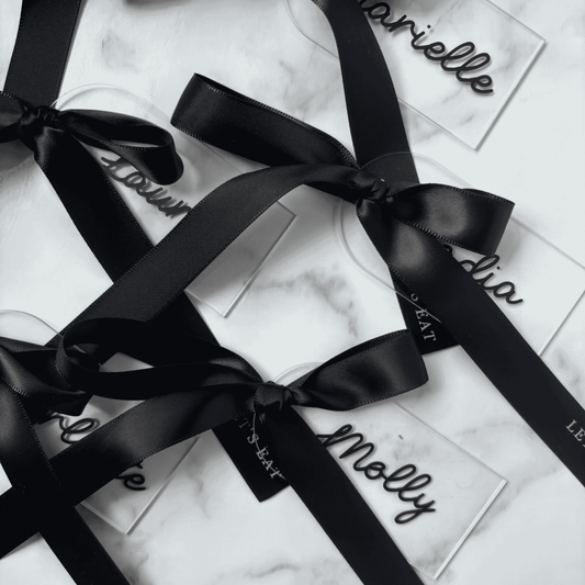 Set of clear acrylic wedding place settings with black names and satin ribbons, styled for modern wedding table settings
