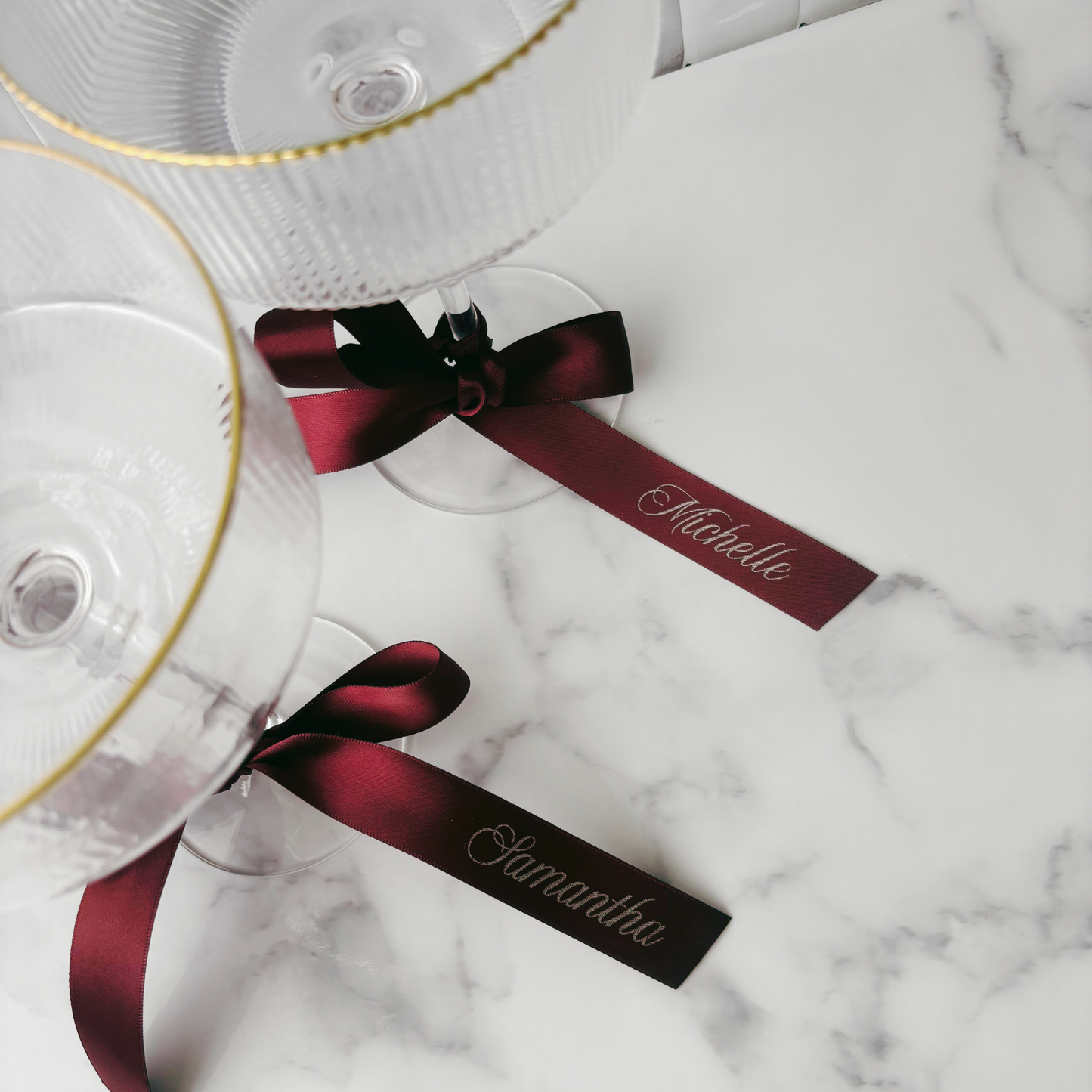 Flat lay of burgundy personalised ribbons tied to coupe glasses with printed guest names