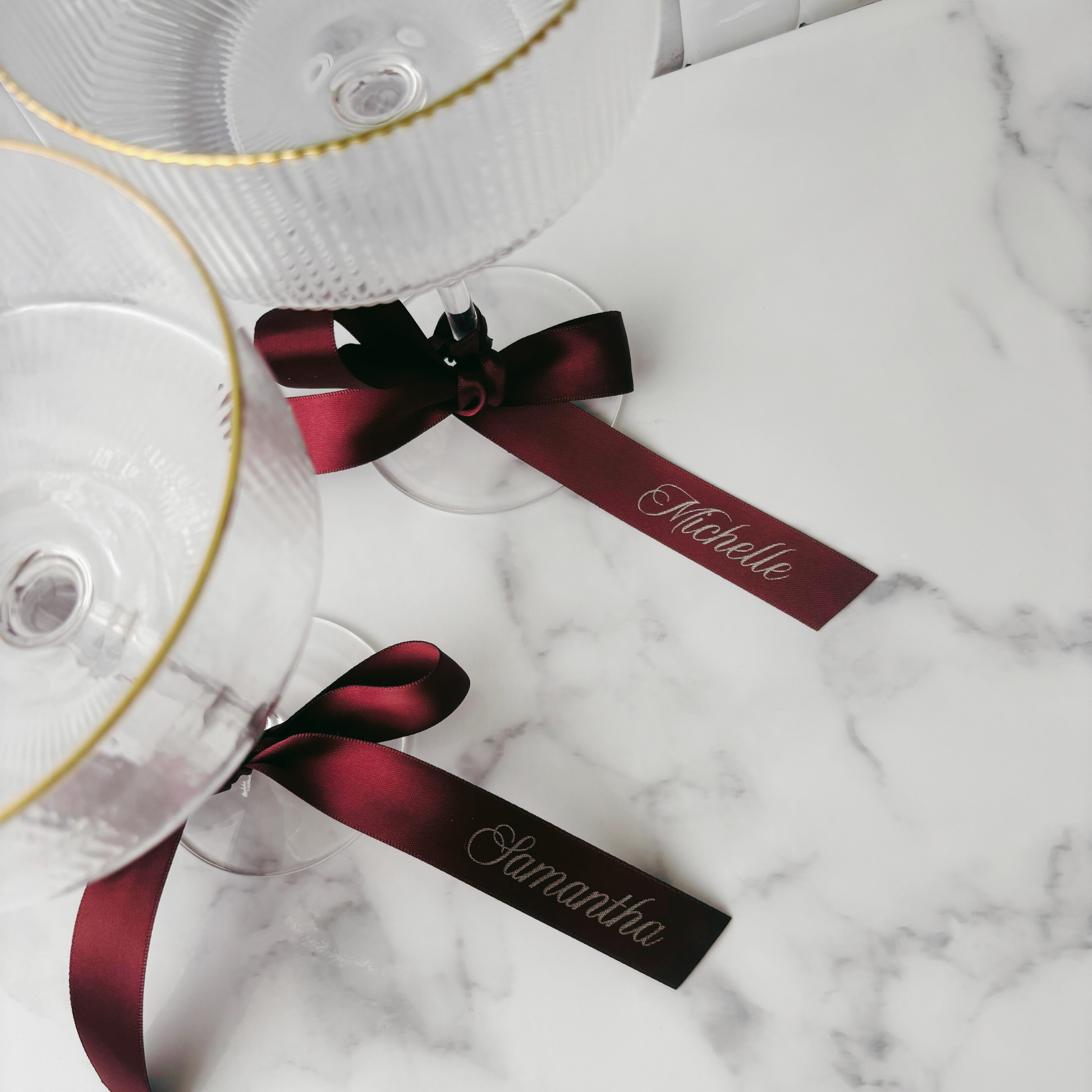 Flat lay of burgundy personalised ribbons tied to coupe glasses with printed guest names