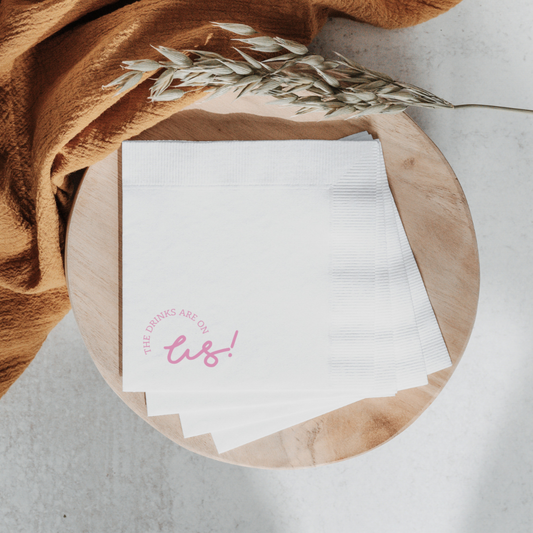 Custom cocktail napkin with pink 'The drinks are on us!' text, perfect for wedding bar signage or party stationery